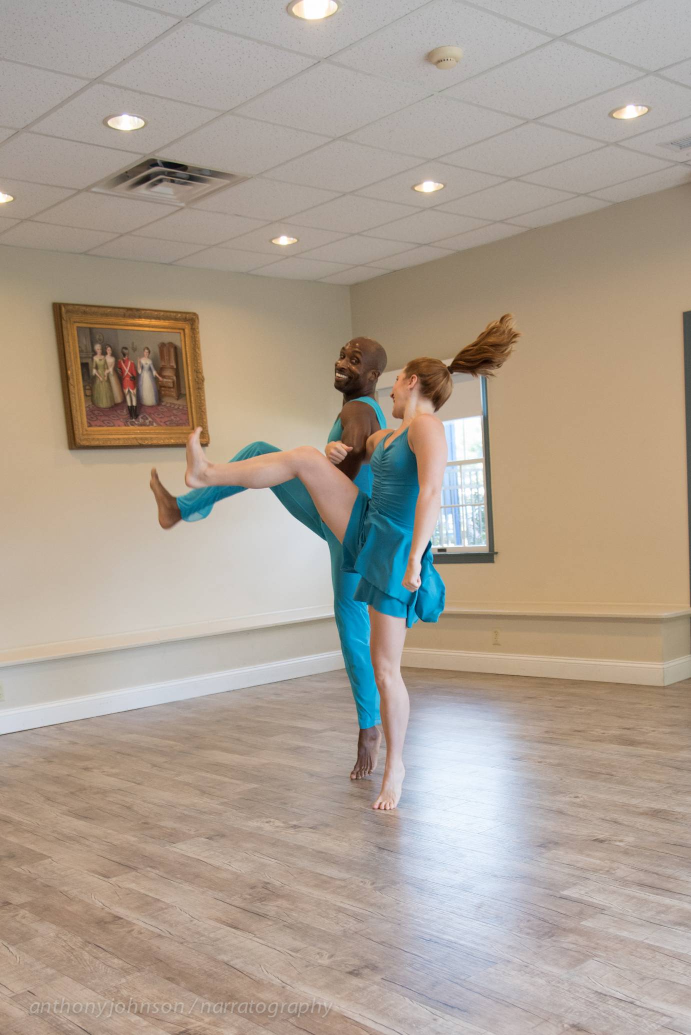 Two dancers perform in a stark gallery space. They wear bright blue costumes as they smile at one another.